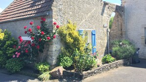 Un edificio de piedra con techo inclinado, un pequeño jardín con flores rojas y amarillas, y una puerta azul.