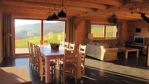 Ein Holzinterieur mit Esstisch, Stühlen, einer Couch, einem Fernseher und großen Fenstern, die einen Blick ins Freie bieten.