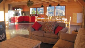 Ein großzügiger Wohnbereich mit Holzboden, einer Couch mit roten Kissen und Blick auf eine Küche und einen Essbereich durch große Fenster.