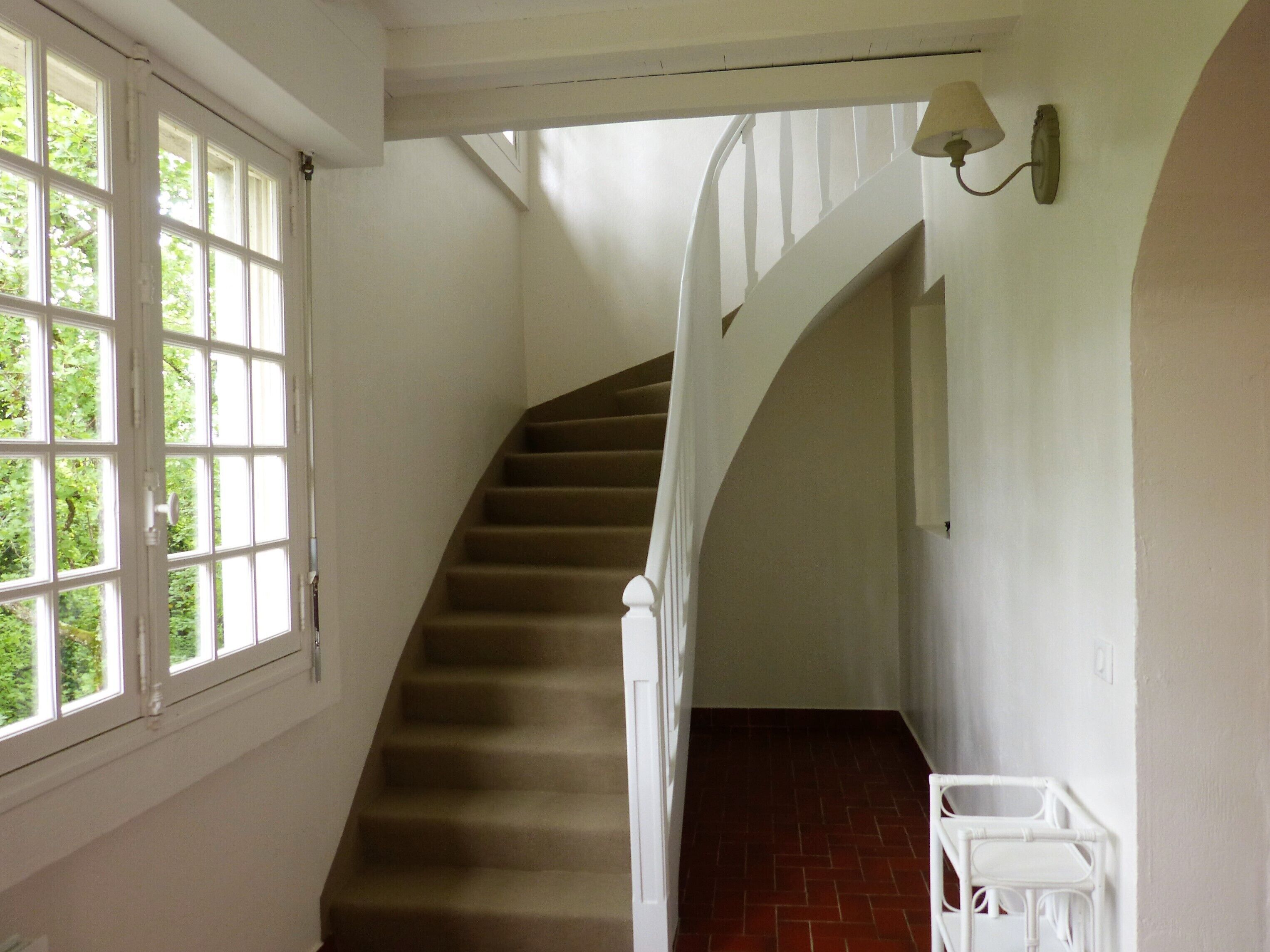 Un escalier avec une rampe en bois et des murs blancs, menant à un palier où se trouvent une chaise blanche et une lampe fixée au mur.