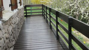 Een houten terras met een stenen muur aan één kant en een zwarte balustrade aan de andere kant, dat leidt naar een gebouw met houten luiken.