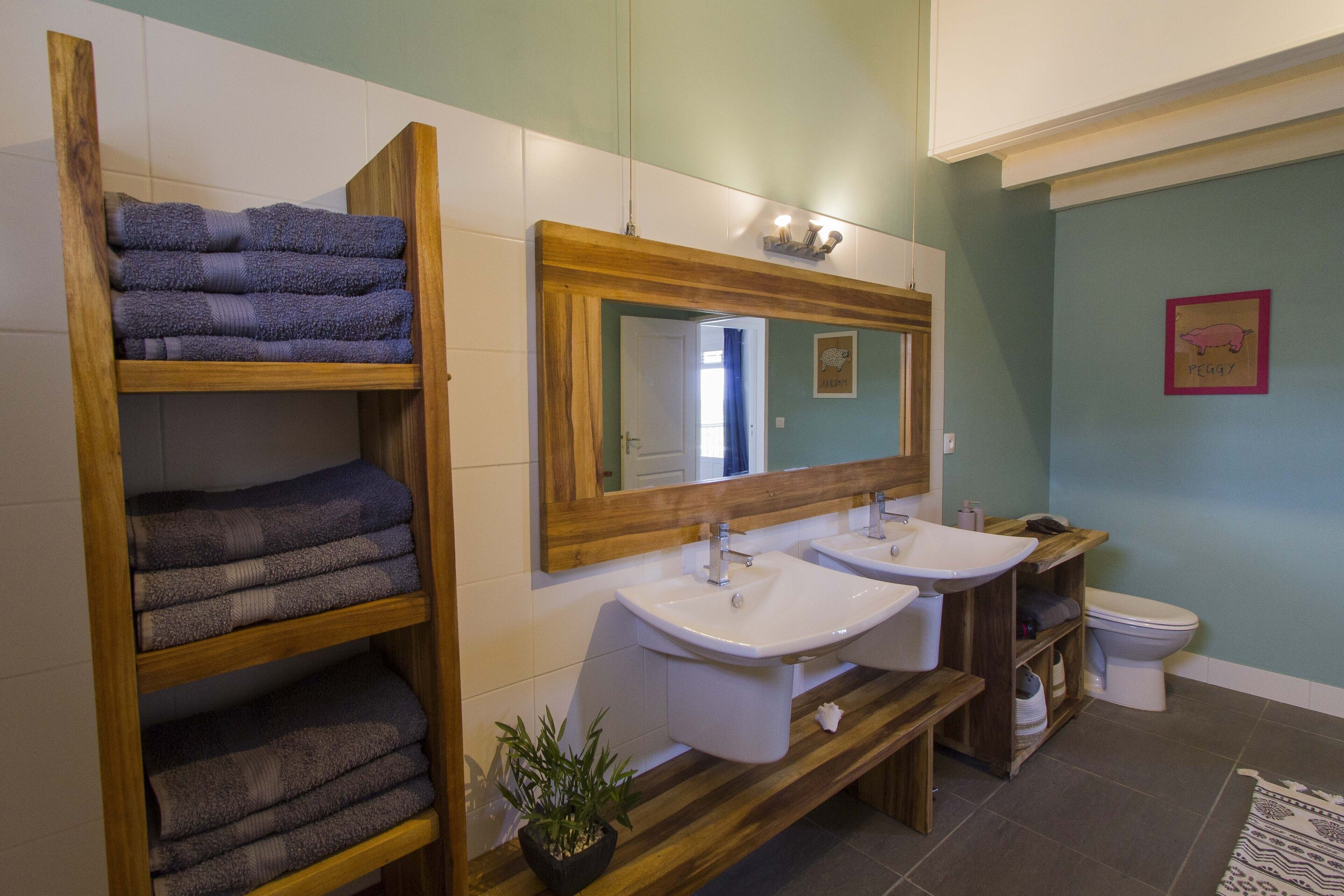 A bathroom with two sinks, a mirror, and a toilet. There are wooden shelves with folded towels.