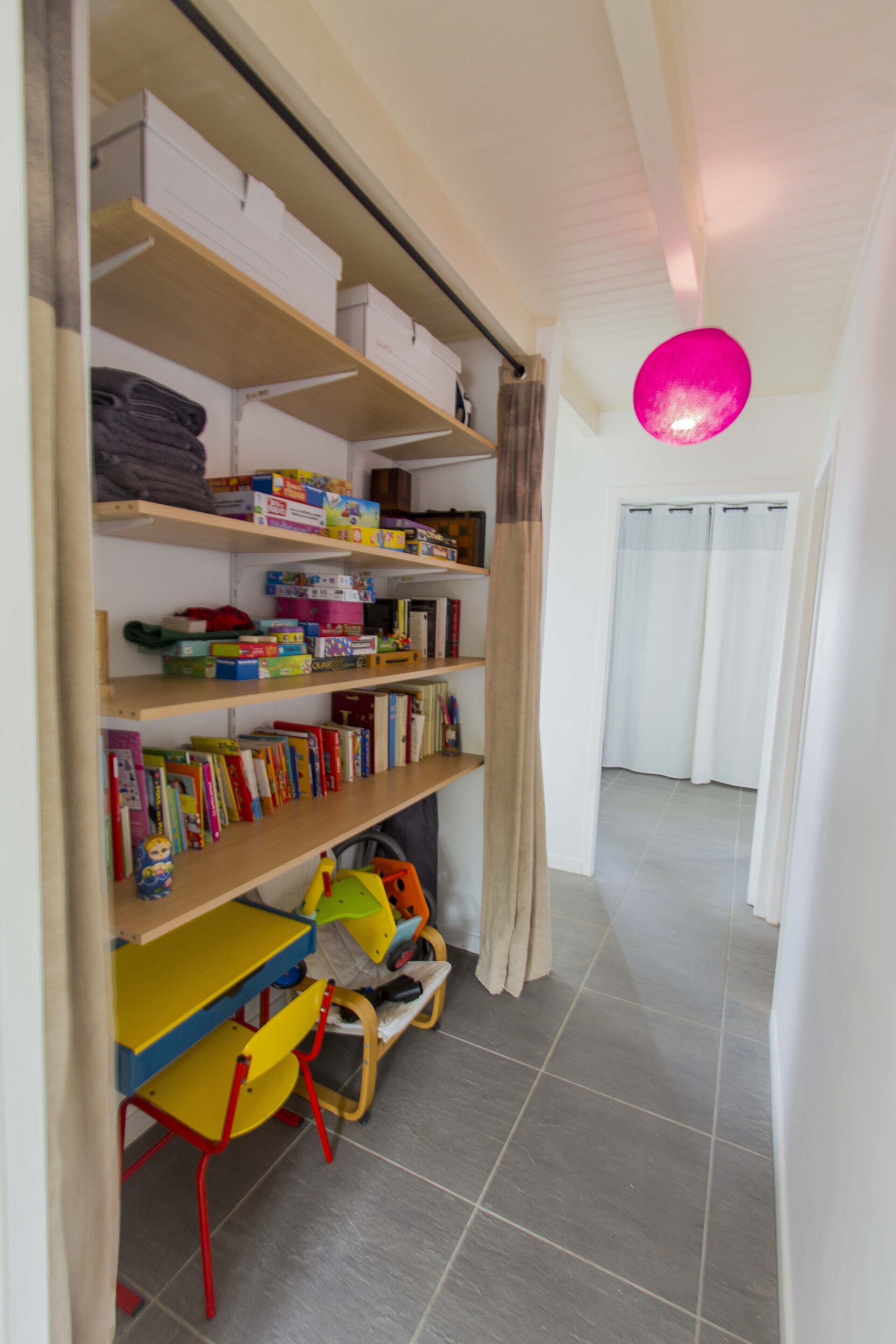 Una habitación con una estantería llena de libros y juguetes, una pequeña mesa con sillas, y una lámpara esférica rosa.