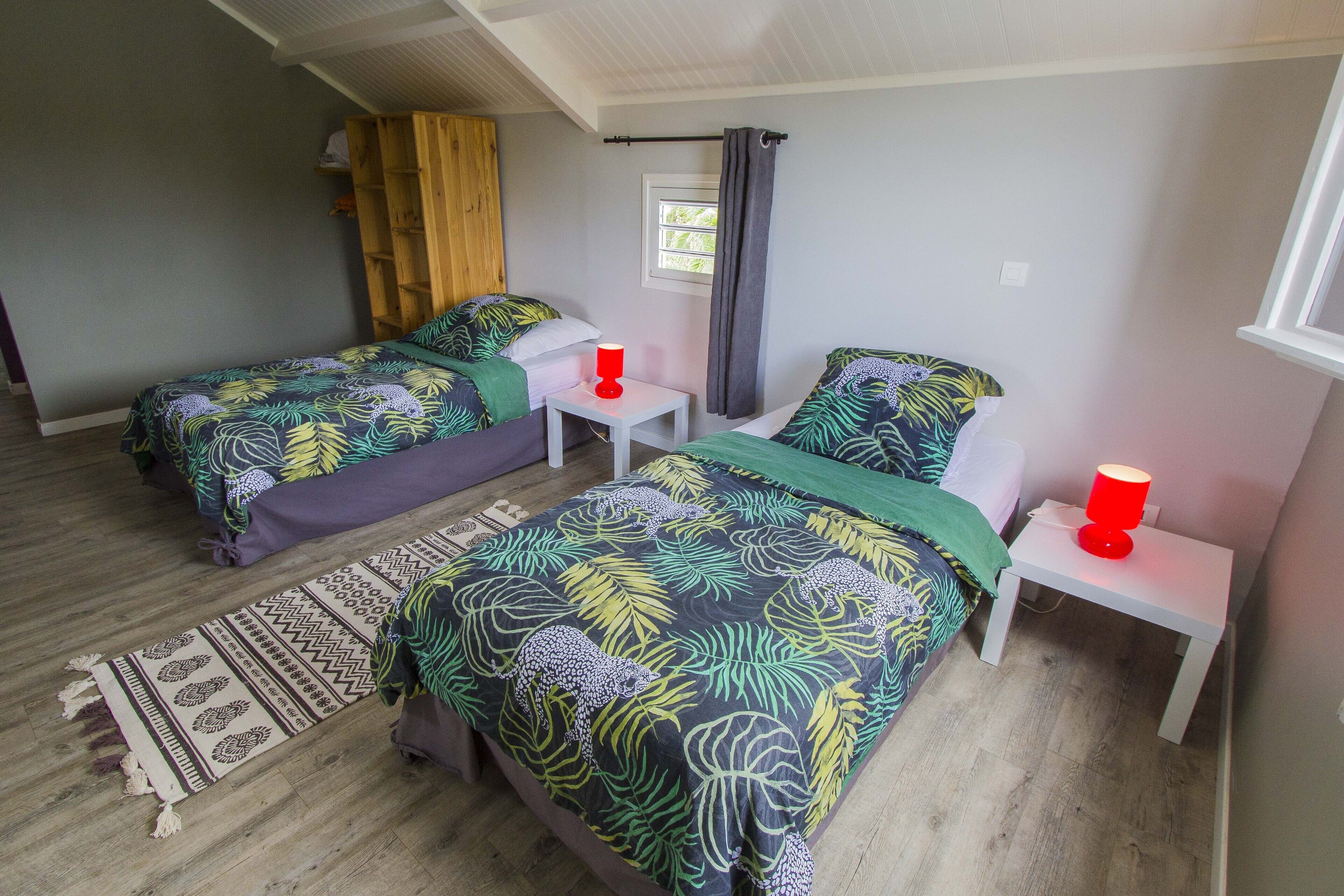 Una habitación con dos camas, cada una con una colcha estampada, una mesita de noche blanca pequeña con una lámpara roja, un estante de madera y una ventana con cortinas.