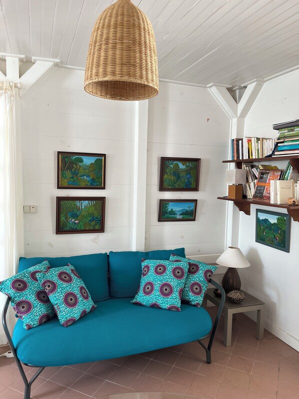 A cozy living room with a teal sofa, patterned cushions, a wicker pendant light, and framed paintings on the walls.