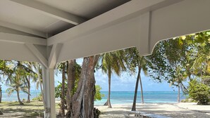 A covered outdoor area with a wooden table and a woven basket, overlooking a sandy beach with palm trees and the ocean. - Large family home with private beach  (Sainte Anne)