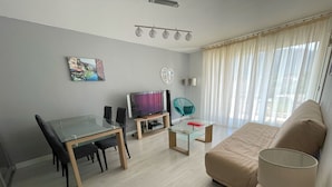 Een moderne woonkamer met een glazen eettafel, een bank, een televisie en een raam met gordijnen.