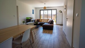 Ein stilvoller Wohnraum mit einem Holzes Esstisch, weißen Stühlen, einer Couch mit Kissen, einem Fernseher und einem großen Fenster.