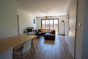 A modern living space with a wooden dining table, white chairs, a sofa with cushions, a television, and a large window.