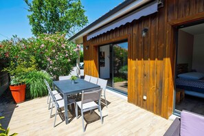 Una terraza de madera con una mesa, sillas, una planta en maceta y un ventanal corredera que da a un dormitorio.