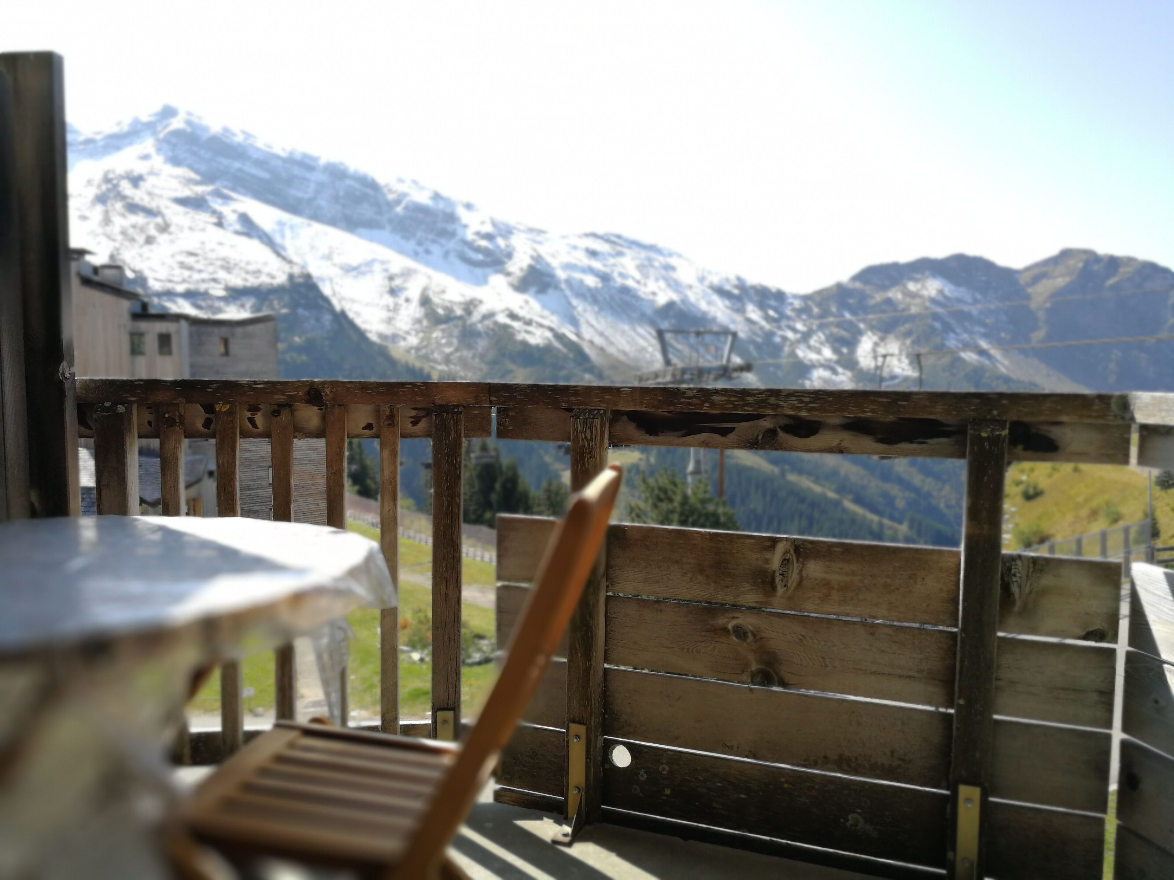 Un balcon en bois avec une table et une chaise, d’où l’on peut admirer des montagnes enneigées.