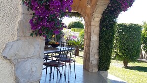 An outdoor patio area with a stone archway covered in purple flowers, a dining table with chairs, and a stone wall. - CHARMING AIR-CONDITIONED VILLA WITH PRIVATE POOL, GARDEN, NEAR THE SEA AND SHOPS (ELS POBLETS)