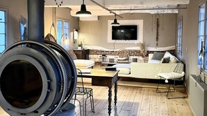 A spacious living room with exposed wooden beams, a large industrial-style heater, a comfortable sofa, and a television mounted on the wall.