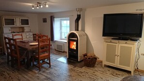 A living room with a fireplace, a dining area with wooden chairs, a television on a cabinet, and a window with curtains.