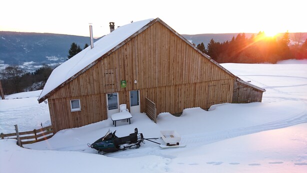 A wooden barn with a snow-covered roof, a snowmobile, and a sled.