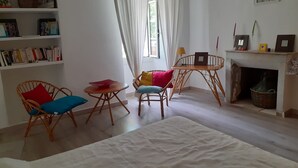 Una habitación acogedora con una estantería, dos sillas de mimbre, una mesita redonda, una cama blanca, una chimenea y un ventanal con cortinas.