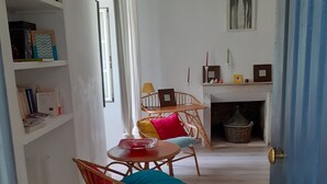 Un estrecho pasillo con sillas de mimbre, una pequeña mesa, una estantería con libros y objetos decorativos, y una chimenea con un cesto.