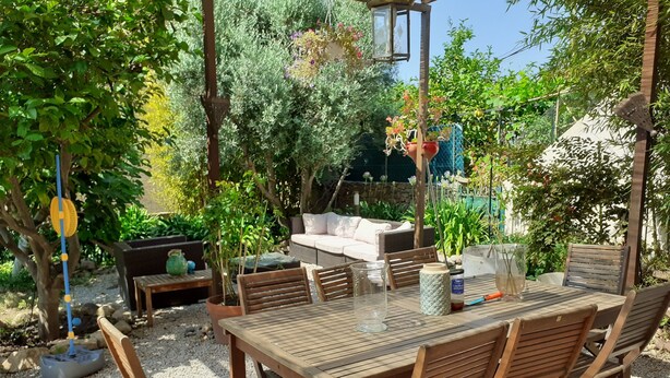 Un patio exterior con mesa y sillas de madera, un sofá y un pequeño jardín con plantas y una fuente.