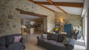 A spacious living area with a stone wall, a wooden ceiling, and a kitchen in the background. - Rental in Nerac en Albret, stone built house, detached, renovated. (Nérac)