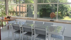 A dining area with a glass table and white chairs, large windows with a view of greenery, and a small table with decorative items.