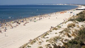 A wide sandy beach with a clear view of the sea, a few boats, and a distant shoreline with buildings. - Apartment with swimming pool - garage - air conditioning. - beach 200m away - WIFI (La Grande-Motte)