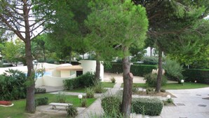 A landscaped garden with various trees and shrubs, a paved walkway, and a building with a curved roof in the background. - Apartment with swimming pool - garage - air conditioning. - beach 200m away - WIFI (La Grande-Motte)