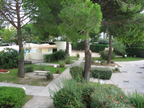A landscaped garden with various trees and shrubs, a paved walkway, and a building with a curved roof in the background. - Apartment with swimming pool - garage - air conditioning. - beach 200m away - WIFI (La Grande-Motte)