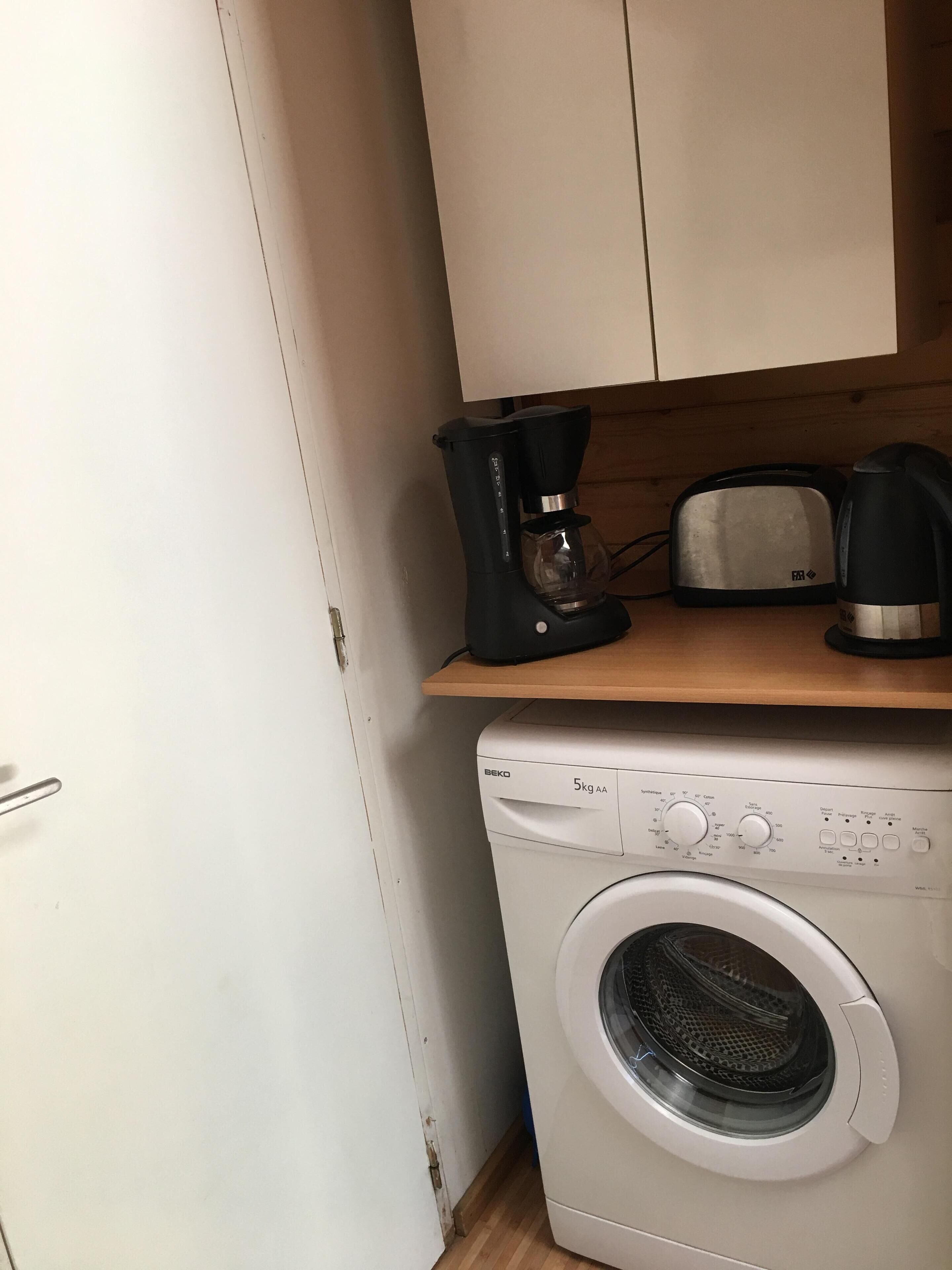 Cocina compacta con lavadora, tostadora y cafetera sobre un estante de madera.