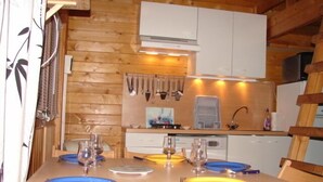 Ein Holzinterieur mit einem für zwei Personen gedecktem Esstisch, einer Küchenzeile mit Schränken und Geschirr sowie einem Fenster mit Vorhängen.