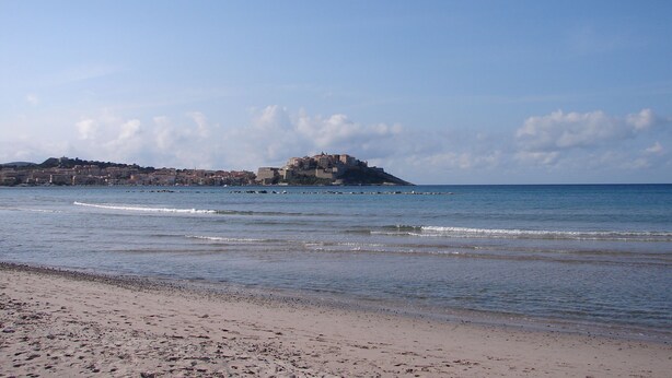 Ein Strand mit einem historischen Gebäude auf einer kleinen Insel, ruhigem Meer und blauem Himmel.