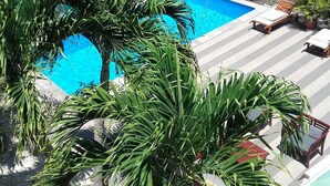 A swimming pool surrounded by lush greenery, a small building with a green roof, and a paved area with lounge chairs.