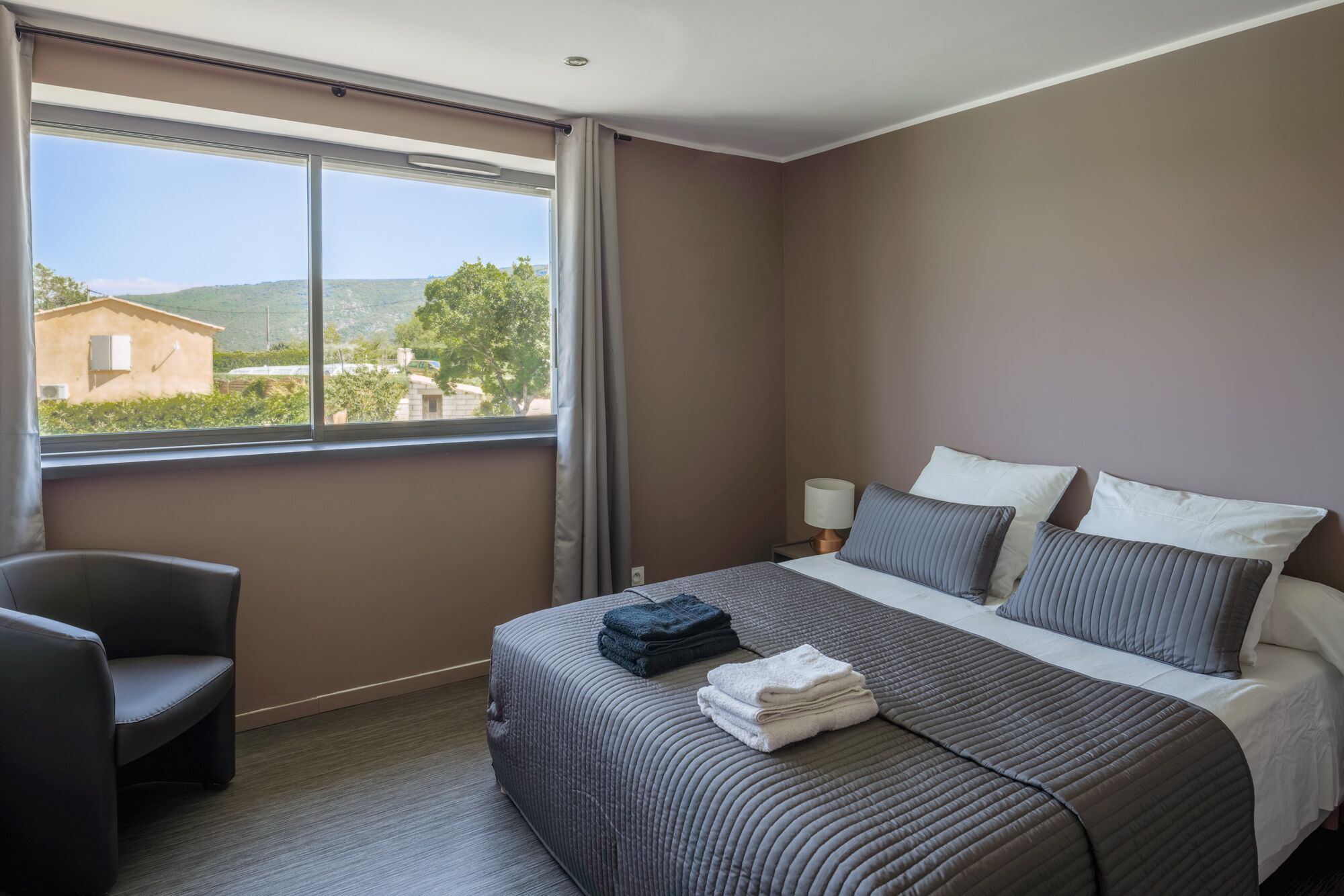 A hotel room with a large bed, a chair, a nightstand with a lamp, and a window with a view of a building and trees.