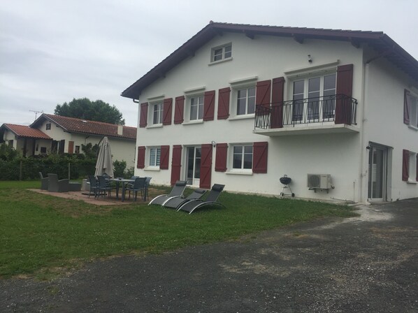 A two-story house with a balcony, a patio with chairs and a table, and a lawn.