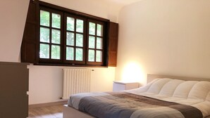 A bedroom with a bed, a nightstand, a window with blinds, and a radiator. - Norman house with pool in the countryside (Bâlines)