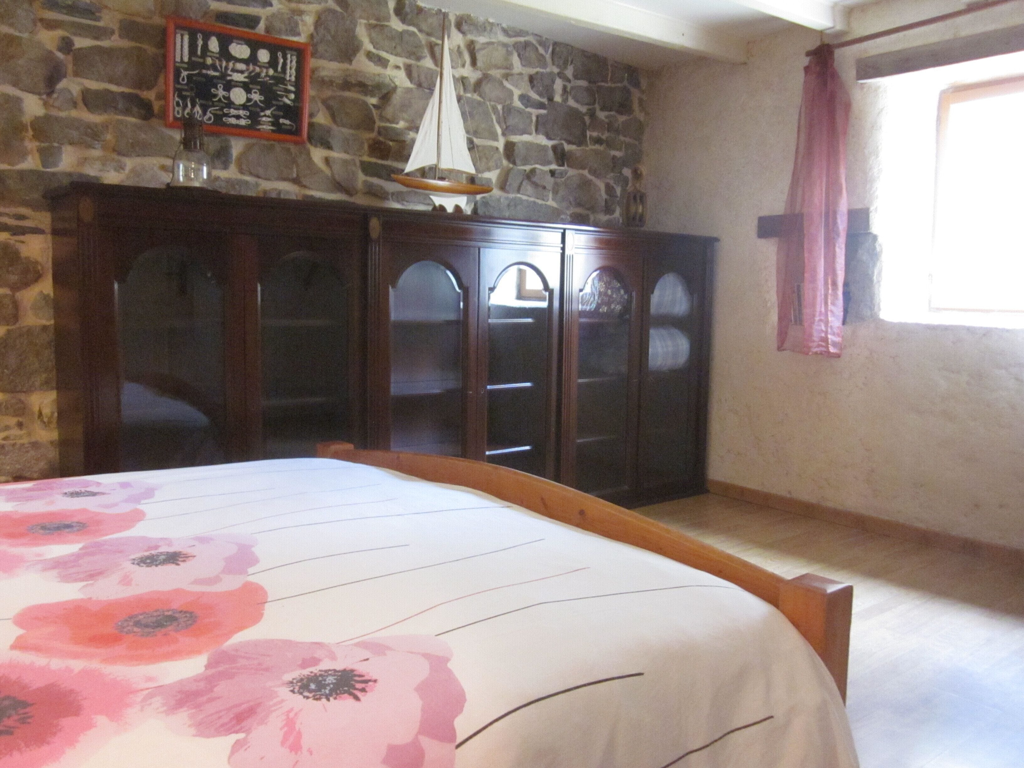 A bedroom with a bed, wooden wardrobe, stone wall, and a window with curtains.