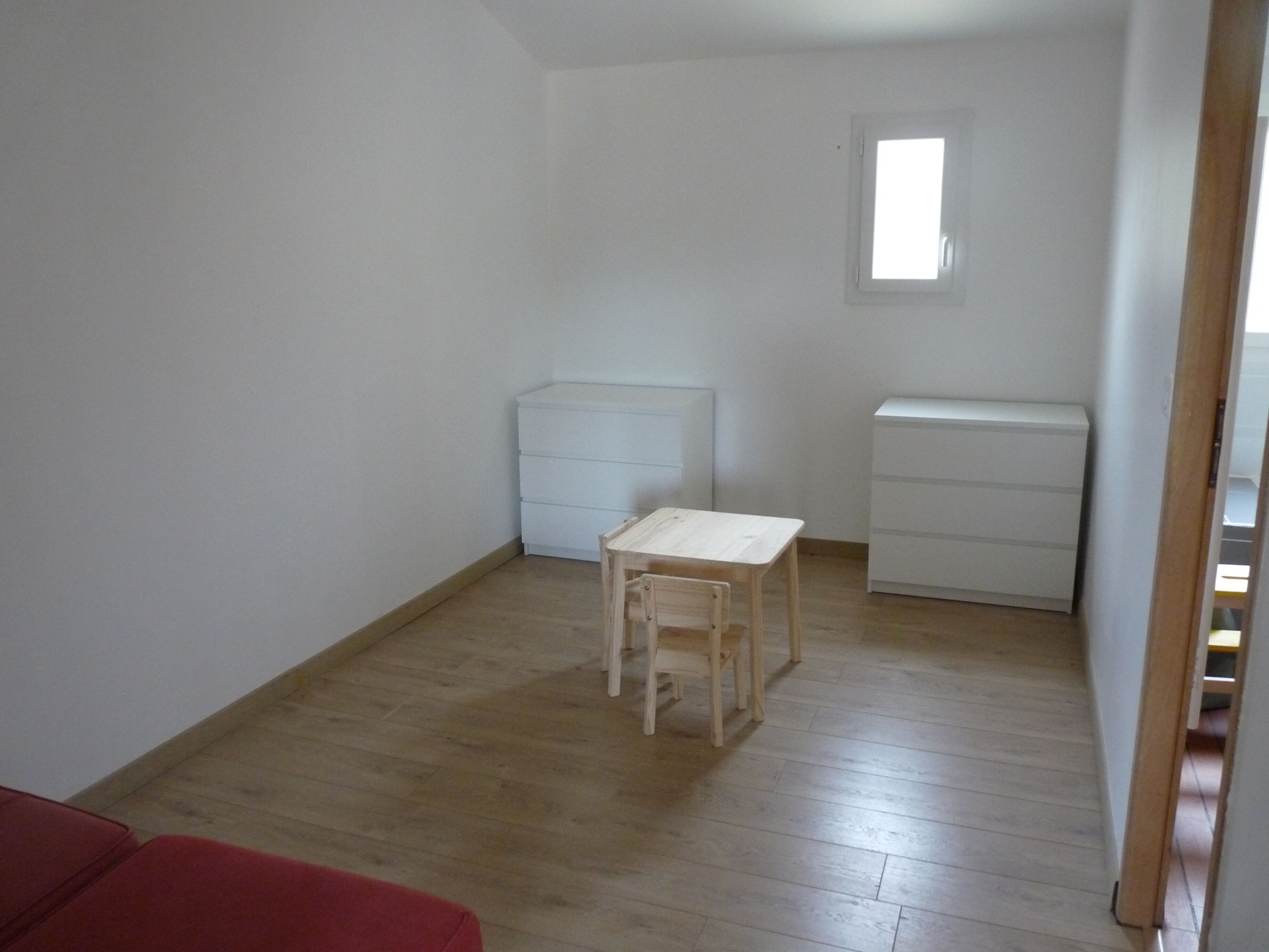 Une petite pièce au mobilier minimaliste, avec un plancher en bois, une petite table, deux commodes blanches et une fenêtre.