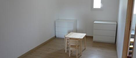 Une petite pièce au mobilier minimaliste, avec un plancher en bois, une petite table, deux commodes blanches et une fenêtre.