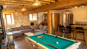 Un salon chaleureux avec une table de billard, une cheminée en pierre et un coin cuisine équipé d’un réfrigérateur et de tabourets de bar.