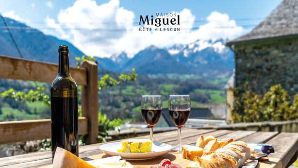 Une table en bois, une bouteille de vin, deux verres de vin rouge, une assiette de fromage, une baguette et un croissant, le tout disposé devant un paysage montagneux pittoresque.