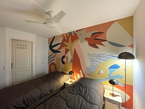 A bedroom with a bed, a desk, a lamp, a ceiling fan, and a colorful abstract wall mural. - La Corniche // 2-room apartment 100 meters from the beach (Sète)