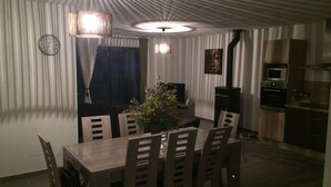 A dining area with a wooden table and chairs, a chandelier, a clock, and a kitchen area with appliances.