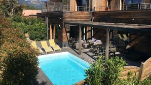 A modern outdoor pool area with wooden deck and lounge chairs, surrounded by greenery and overlooking a mountainous landscape.