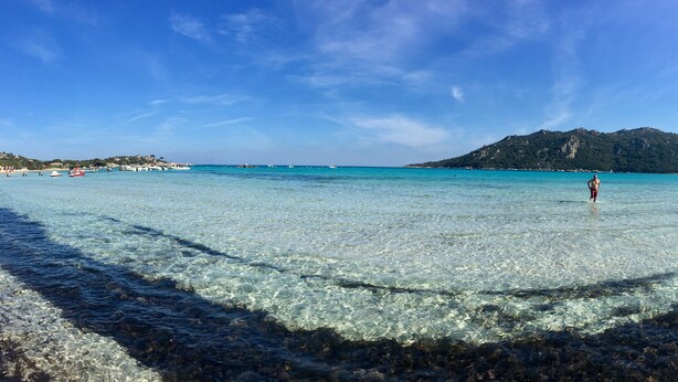 A beach with clear shallow waters, a person standing in the water, and a mountain in the background.