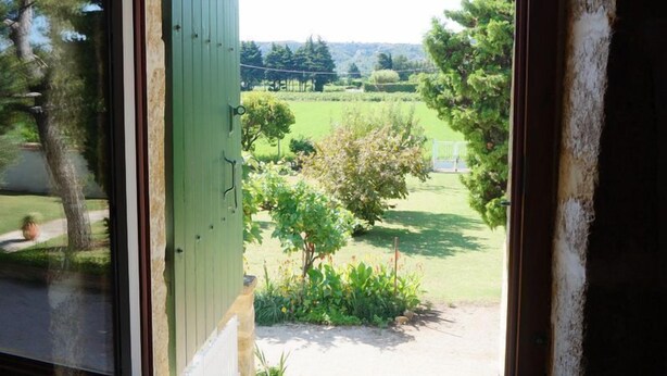 A view through a window with a wooden frame, revealing a green door and a garden with trees and a lawn. - Gîte in an early 18th-century Provencal farmhouse (Roquemaure)