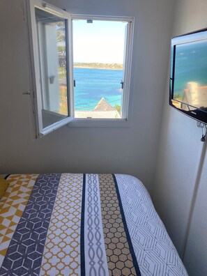 A bedroom with a bed, a window with an open view of the sea, and a wall-mounted TV. - PROMO - Duplex PANORAMIC SEA VIEW, Quiet and close to the BEACH  (Perros-Guirec)