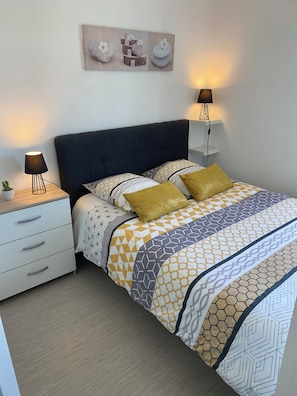 Une chambre à coucher bien rangée, avec un lit, des tables de chevet, une petite plante et une œuvre d’art accrochée au mur.