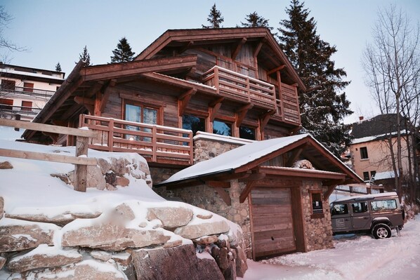 A wooden chalet with a stone foundation, a balcony, and a vehicle parked nearby.