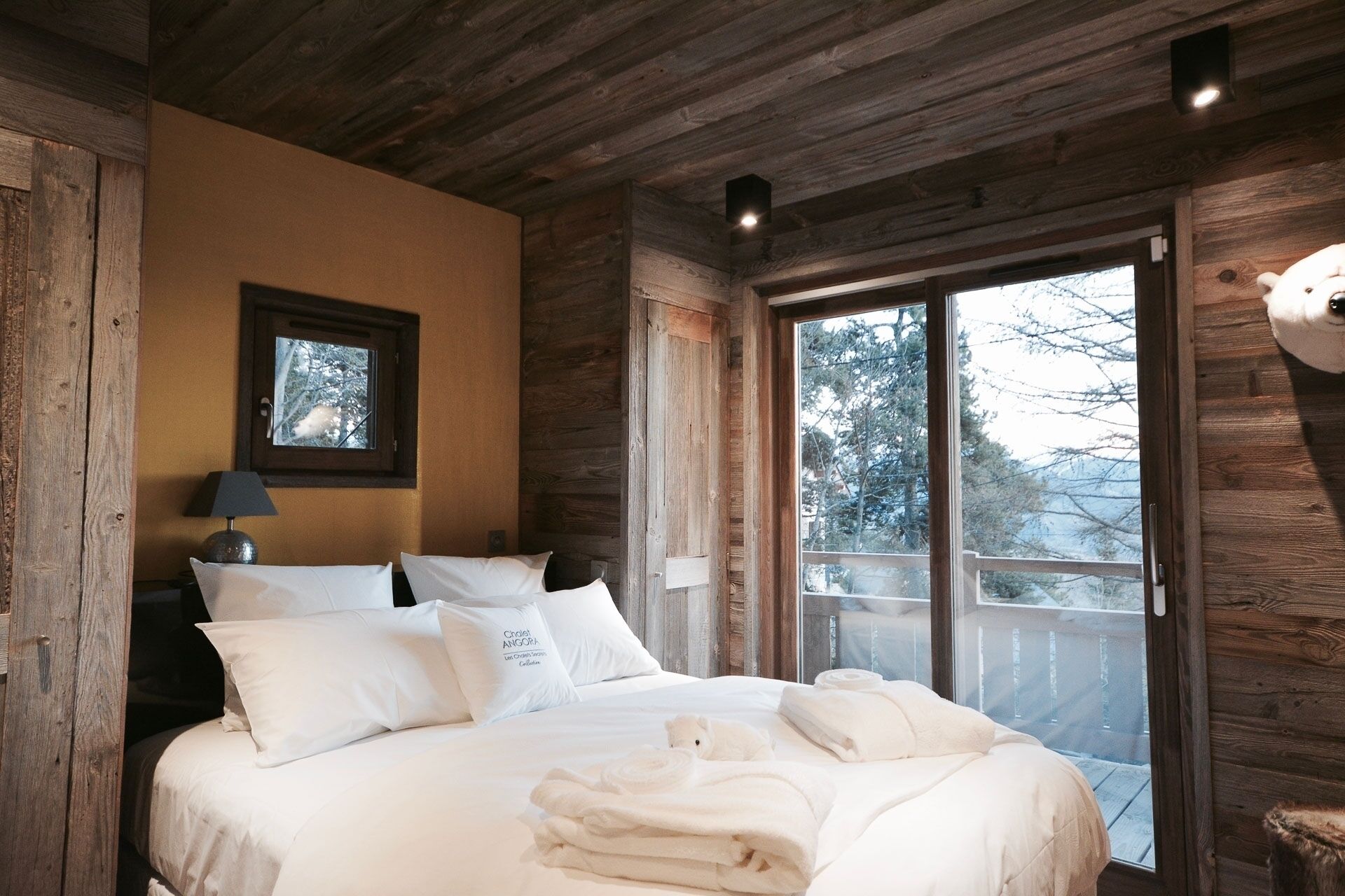 Une chambre à coucher dans une cabane en bois, avec un lit, des oreillers, une table de chevet, une lampe et une fenêtre donnant sur la forêt.
