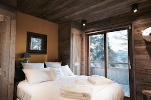 Un dormitorio de cabaña de madera con cama, almohadas, una mesita de noche, una lámpara y una ventana con vistas a árboles.