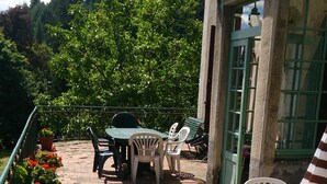 An outdoor patio area with a brick floor, green and white plastic chairs, a wooden table, and potted plants. - Large family house sleeps 16 in the heart of a beautiful medieval village (Génolhac)